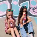Twee meisjes zitten en drinken uit rode Kambukka drinkflessen met vogels en regenboog, zittend voor graffiti.