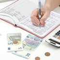 Hand vult een budget planner in met een pen, met eurobiljetten, munten en een rekenmachine op tafel.