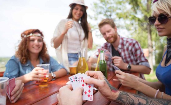 Vijf vrienden spelen kaarten aan een picknicktafel in de buitenlucht, met bier en drankjes.