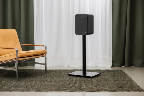 Zwarte Ultima speaker op een zwarte staander in een woonkamer met stoel en gordijnen.