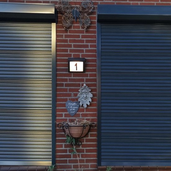 Klein zwart huisnummerbord met verlichting, nummer '1'. Tussen twee rolluiken, met decoratie vlinder en gezicht.