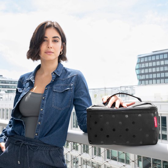 Vrouw met sproeten, gekleed in een spijkerblouse, houdt een zwarte toilettas met stippen vast.