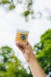 Een hand houdt een potje Blije Voeten eelt- en klovencrème vast, met het label naar voren gericht. De achtergrond is wazig groen gebladerte.