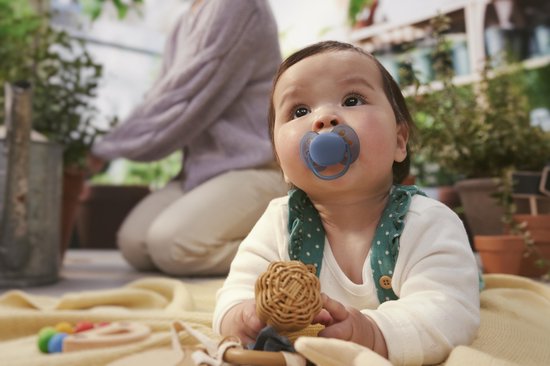 Baby met blauwe Philips Avent fopspeen in de mond, zittend op een gele deken buiten.