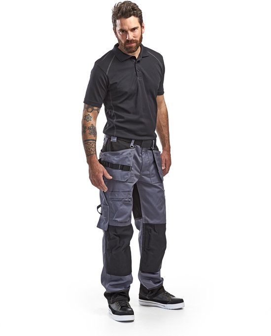 Homme en pantalon de travail gris, chemise noire, avec poches à outils.