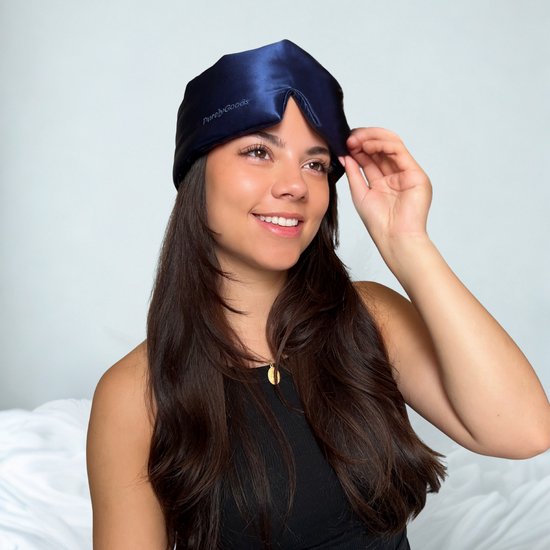 Jeune femme aux longs cheveux bruns, portant un masque de sommeil en soie bleu marine et souriant.