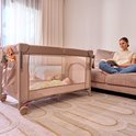 Een vrouw leest een boek naast een beige reisbedje met een slapende baby erin. Het reisbedje is bevestigd aan een ouderlijk bed.