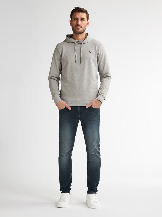 Man met handen in zakken, draagt grijze hoodie en donkerblauwe jeans.