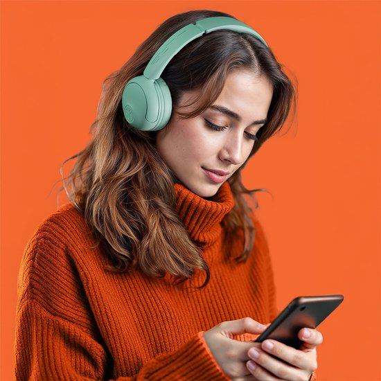 Vrouw met bruin haar in een oranje coltrui draagt een mintgroene JLab Studio 2 koptelefoon en kijkt naar haar telefoon.
