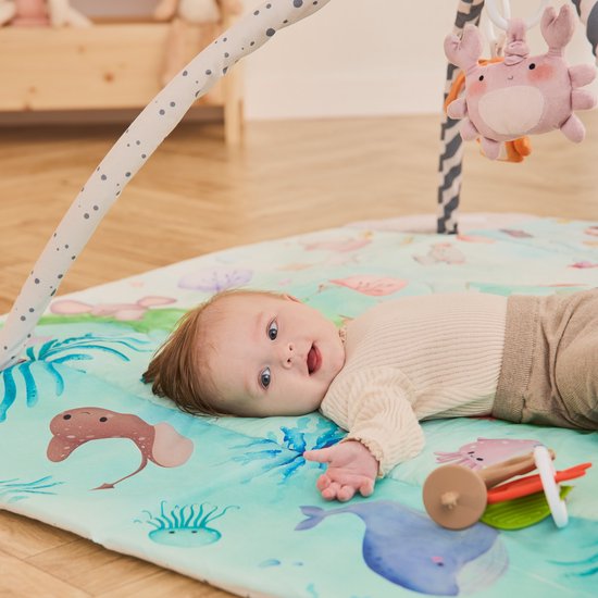 Baby ligt op een speelkleed met waterdieren en planten, met een roze krab boven zich.