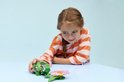 Een meisje met een vlecht, een oranje gestreepte trui en een roze broek, speelt met een groene legoschildpad en bloem.