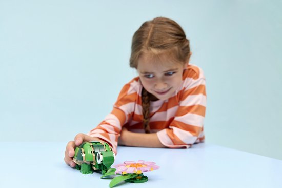 Een meisje met een vlecht, een oranje gestreepte trui en een roze broek, speelt met een groene legoschildpad en bloem.