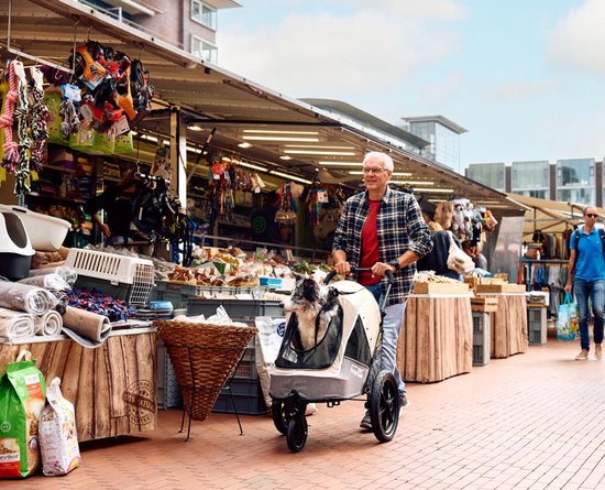 Man duwt beige en grijze hondenbuggy met hond op een markt.