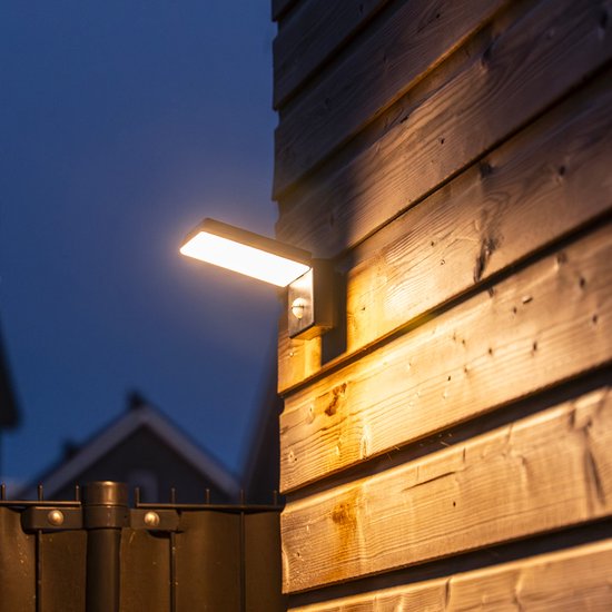 Zwarte wandlamp met sensor, verlicht de houten schutting met warm wit licht.