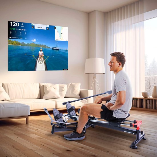 Man op een inklapbare zwarte roeitrainer in een woonkamer, kijkend naar een virtuele roeisimulatie.