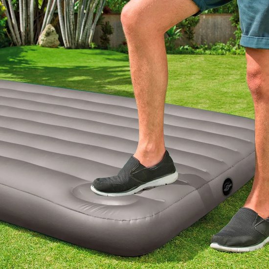 Een man stapt met zijn rechtervoet op een grijs Intex luchtbed in een grasrijke tuin.