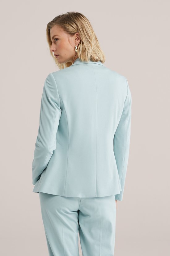 Lichtblauwe blazer met lange mouwen, gedragen door een vrouw met haar handen in de zakken, rug zichtbaar.