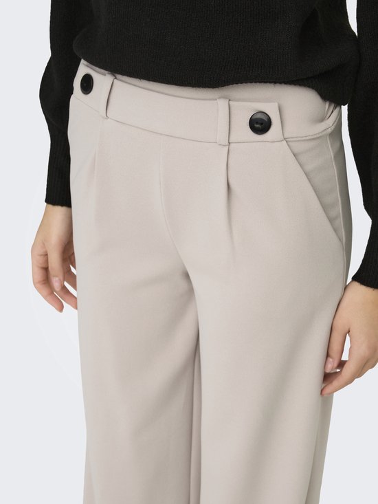 Detail van een lichtbeige wijde broek met plooitjes en twee zwarte knopen aan de voorkant.