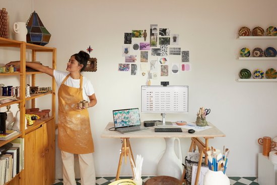Une femme en tablier jaune prend de la céramique sur une étagère dans un studio avec de l'art mural, un ordinateur portable et un clavier HP sur un bureau.