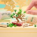Miniatuur bonsai boom met bruine stam en groene bladeren, omringd door steentjes en zakjes.