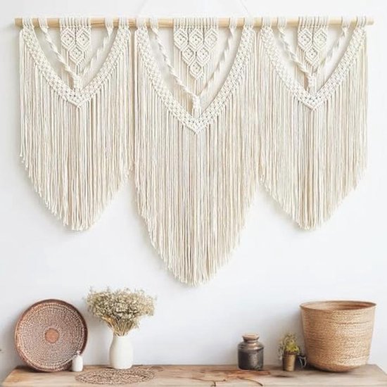 Crèmekleurig macramé wandkleed met franjes boven een houten meubel met manden en een vaas met droogbloemen.