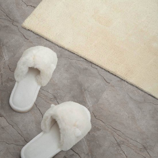 Witte pluizige slippers liggen op grijze tegels naast een crèmekleurig badmat.