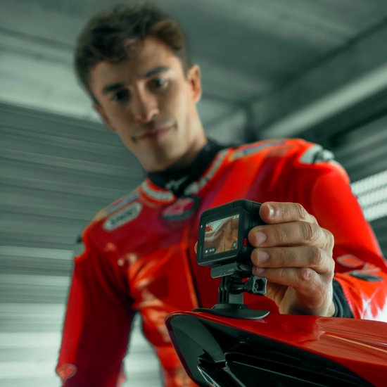 Marc Márquez in racepak houdt een Insta360 actiecamera omhoog voor een rode motorfiets.