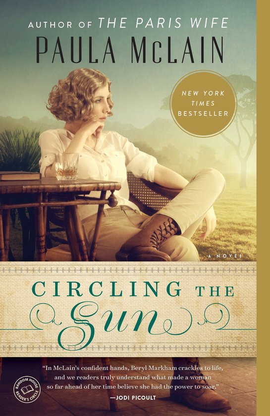 Boek "Circling the Sun" van Paula McLain. Een vrouw met kort, golvend haar zit ontspannen op een stoel met rieten zitting, met een glas op tafel.