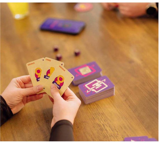 Een persoon houdt kaarten van "Music Match" vast met tekst zoals "VOCALS" en "LAND" in handen. Op tafel liggen paarse stapels kaarten.
