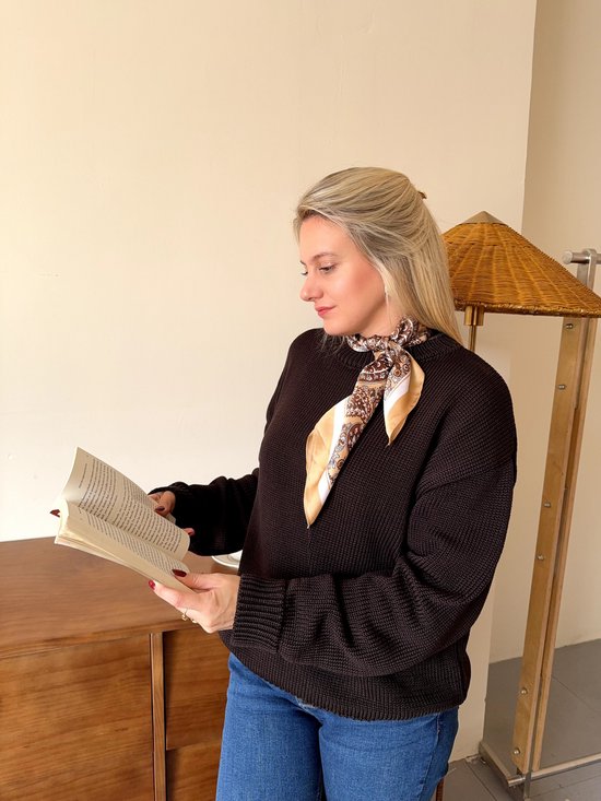 Model draagt een bruine trui, blauwe jeans en een crème sjaal met paisley-print om de nek, leest een boek.