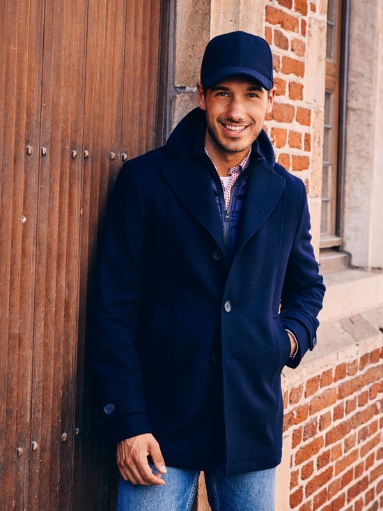 Man met blauwe pet en cabanjas, draagt geruite blouse en blauw gilet, leunt tegen stenen muur.