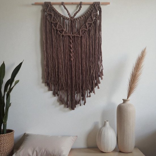 Bruin macramé wandkleed met franjes, aan een houten stok, hangend aan een muur met planten en vazen.