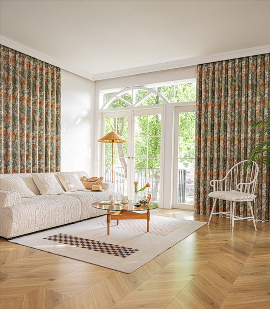 Woonkamer met een bank, salontafel en een groot raam met groen gordijn met bloemenprint.
