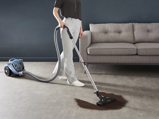 Femme aspirante une tache sur un tapis gris avec un aspirateur Rowenta bleu.
