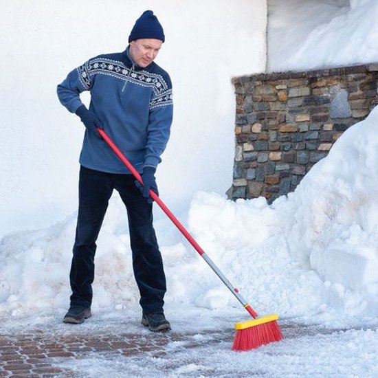 Man met blauwe trui en muts veegt sneeuw van terras met rode bezem met gele borstels.