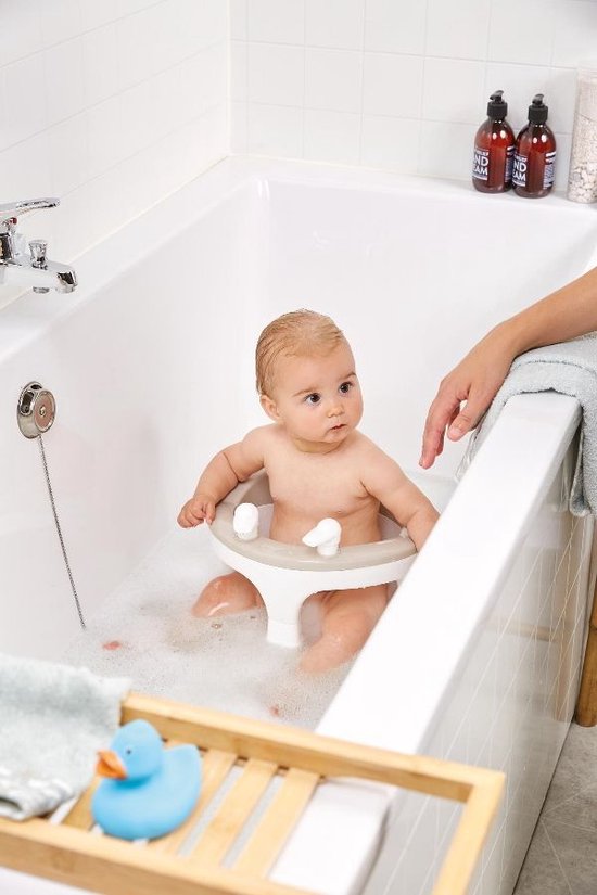Baby zit in het Bébé-jou badzitje in een bad met bubbels, een hand raakt de rand aan.