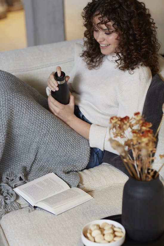 Vrouw op een bank, onder een deken, drukt op de rode dop van een zwarte Tefal Bludrop thermosfles.
