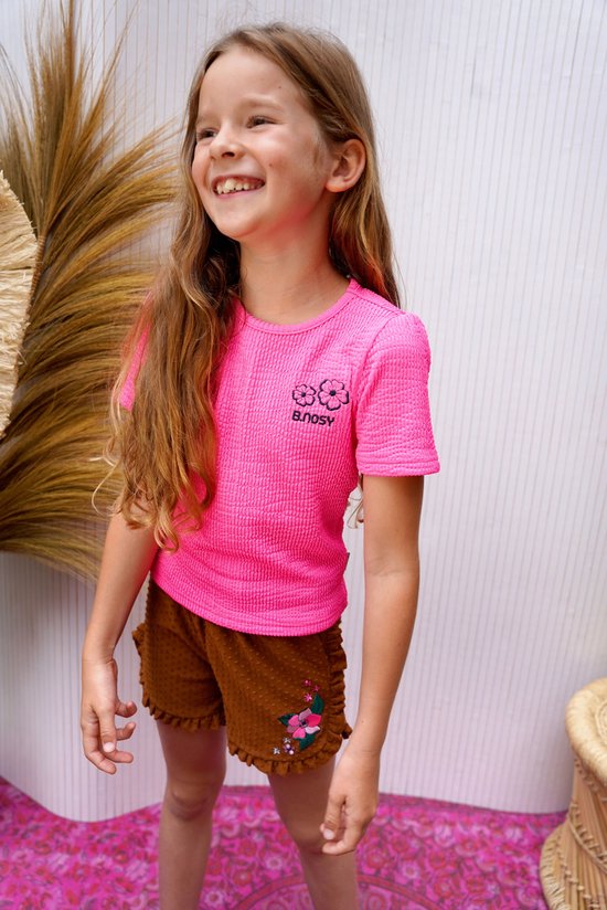 Een lachend meisje met lang blond haar draagt een felroze top met structuur en korte mouwen met bloemenprint en 'B. Nosy'.
