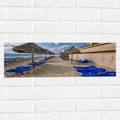 WallClassics - Muursticker - Blauwe Ligbedden op Strand met Rieten Parasols - 60x20 cm Foto op Muursticker