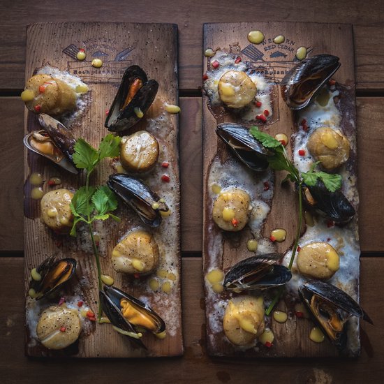 Twee cederhouten planken met sint-jakobsschelpen en mosselen, besprenkeld met saus en rode peper.