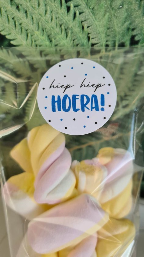 Een doorzichtige zak met pastelkleurige (roze, gele, witte) gedraaide marshmallows. Op de zak zit een ronde blauwe sticker met de tekst 'hiep hiep HOERA!'.