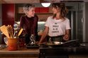 Twee vrouwen in een keuken; één met kort blond haar in een bordeauxrode coltrui, de ander met bruin haar draagt een wit T-shirt met 'WE SHOULD ALL BE FEMINISTS'.