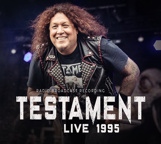 Zanger Chuck Billy van Testament lacht op een podium, draagt een zwart T-shirt met "TAME" erop, een zwarte leren jas met studs en lange krullende bruine haren.