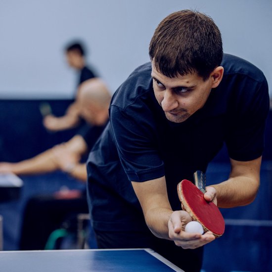 Man met zwarte polo die een tafeltennisbal vasthoudt in zijn hand, klaar om te serveren met een rood tafeltennisbat.