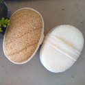 Twee ovale douche sponzen, één van natuurlijk loofah en één van wit badstof, op een grijze ondergrond.