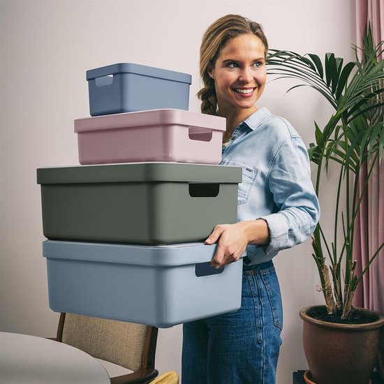 Vrouw met een staart, lachend en een stapel van 4 gekleurde opbergdozen houdend. Van onder naar boven: lichtblauw, roze, groen, lichtblauw.