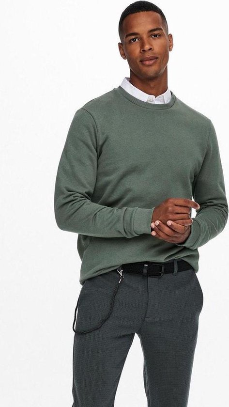 Man kijkt naar de zijkant, handen tegen elkaar. Draagt een groene sweater, wit overhemd en grijze geruite broek met riem.