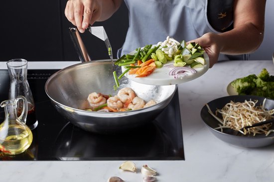 Een wokpan met garnalen en olie op een kookplaat, er worden groenten aan toegevoegd.