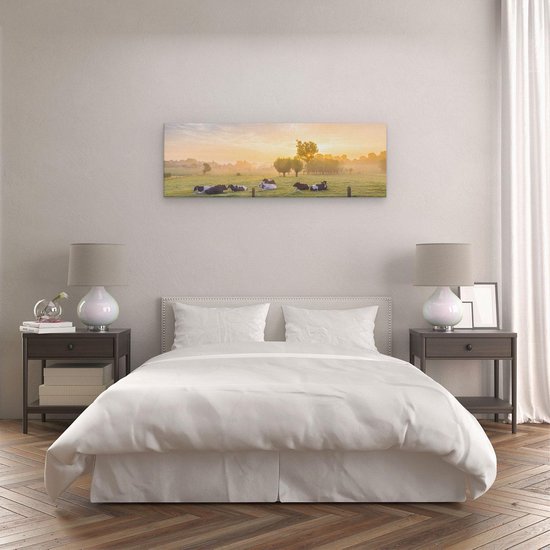 Panorama canvas van koeien in de wei bij zonsopgang, hangend boven een wit bed in een slaapkamer met bruine bijzettafels.