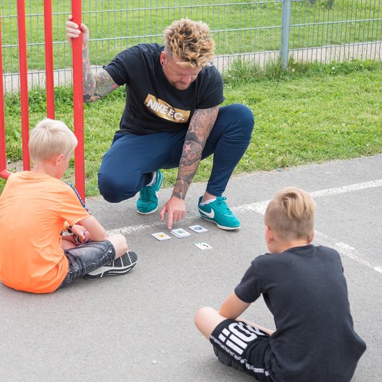 Man met tatoeages wijst naar kaarten op de grond, zittend met twee jongens op een sportveld.